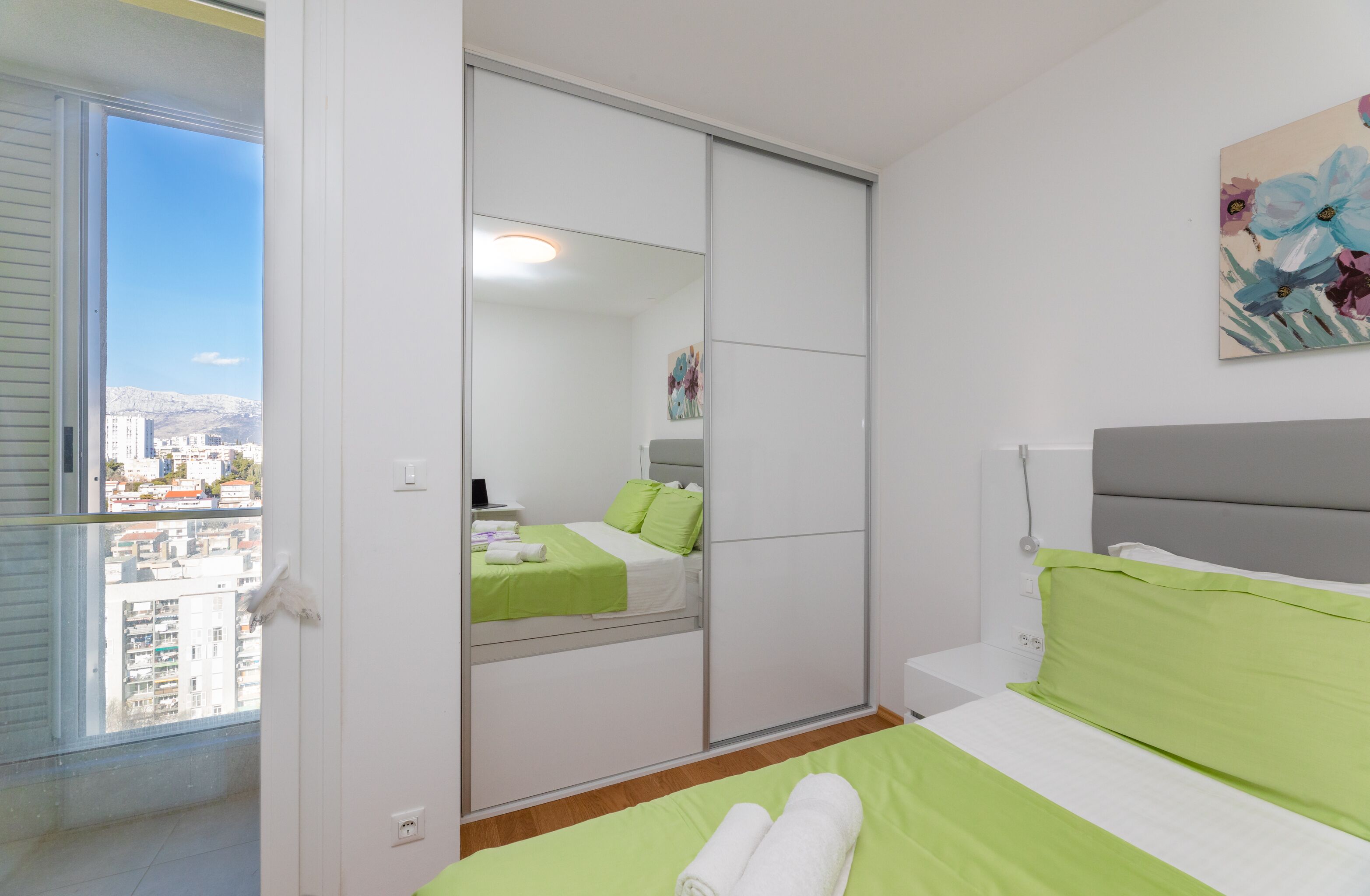 Una camera da letto con un letto, biancheria verde, una porta scorrevole per l'armadio, un quadro e vista sulla città dalla finestra.
