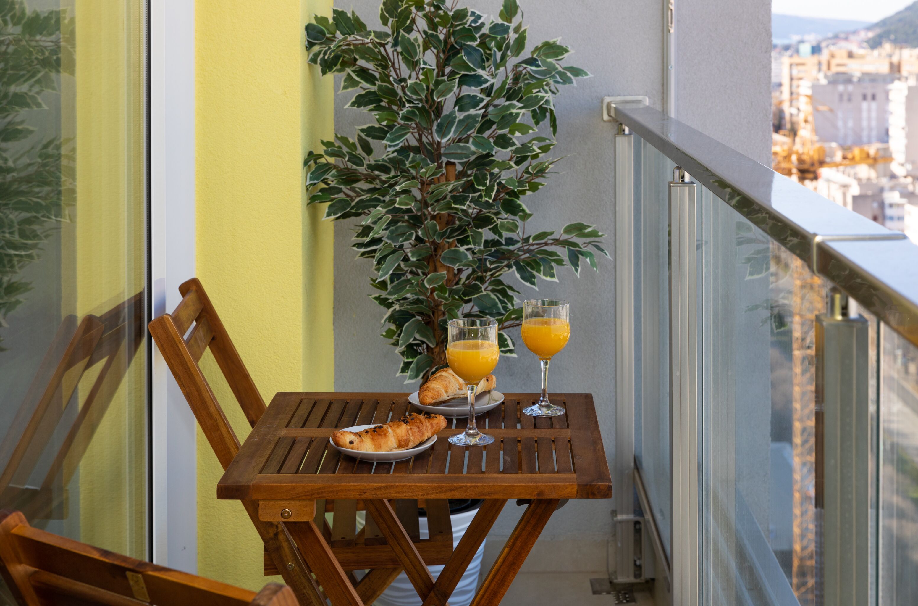 Ein Balkon mit einem Holztisch, zwei Orangensaftgläsern, einem Tablett mit Croissants und einer Topfpflanze.
