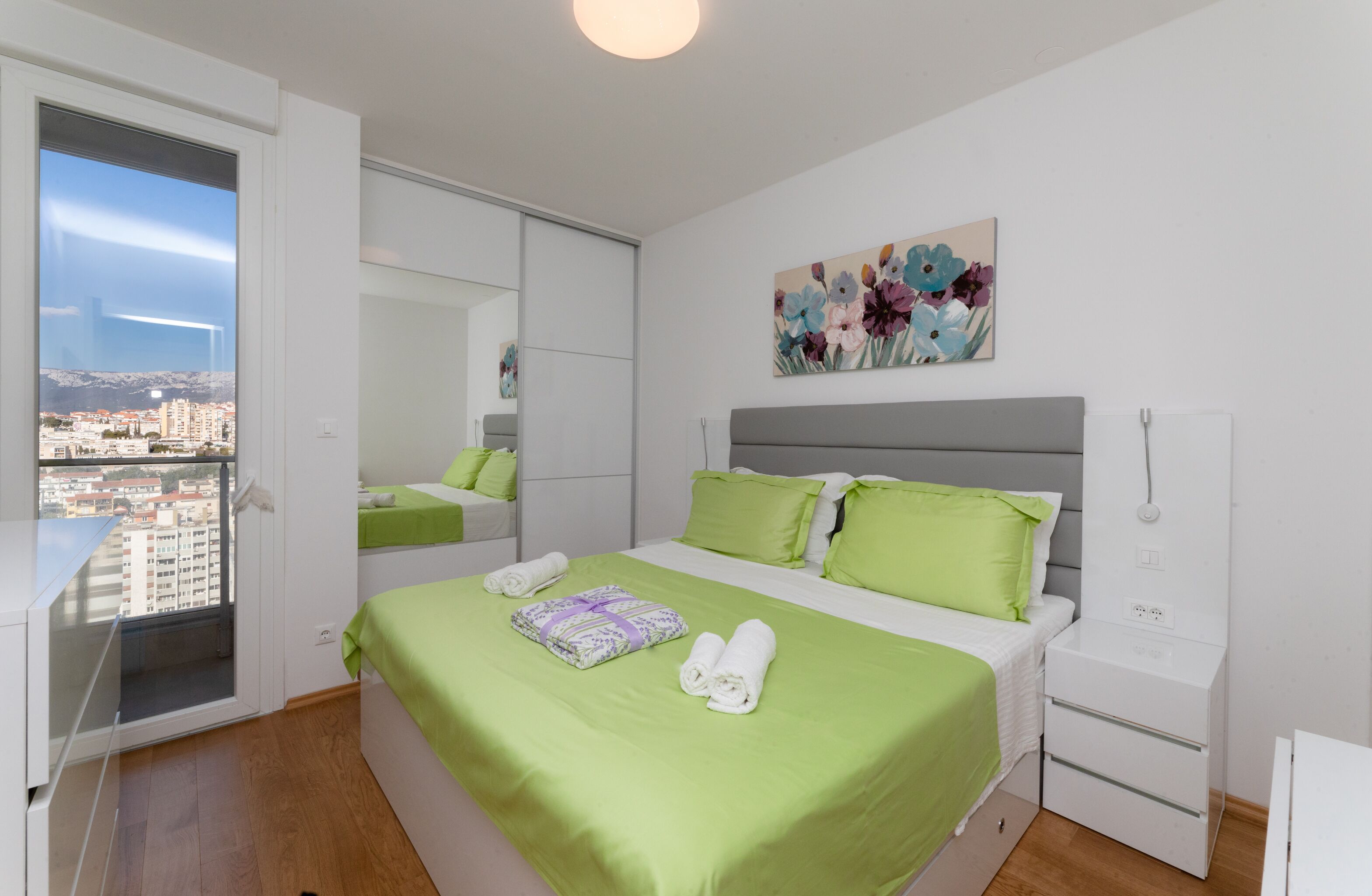 A bedroom with a bed, green bedding, a nightstand, a painting, and a view of a city through a window.