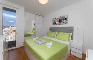 Quarto com cama, roupa de cama verde, mesa de cabeceira, pintura e vista da cidade pela janela.