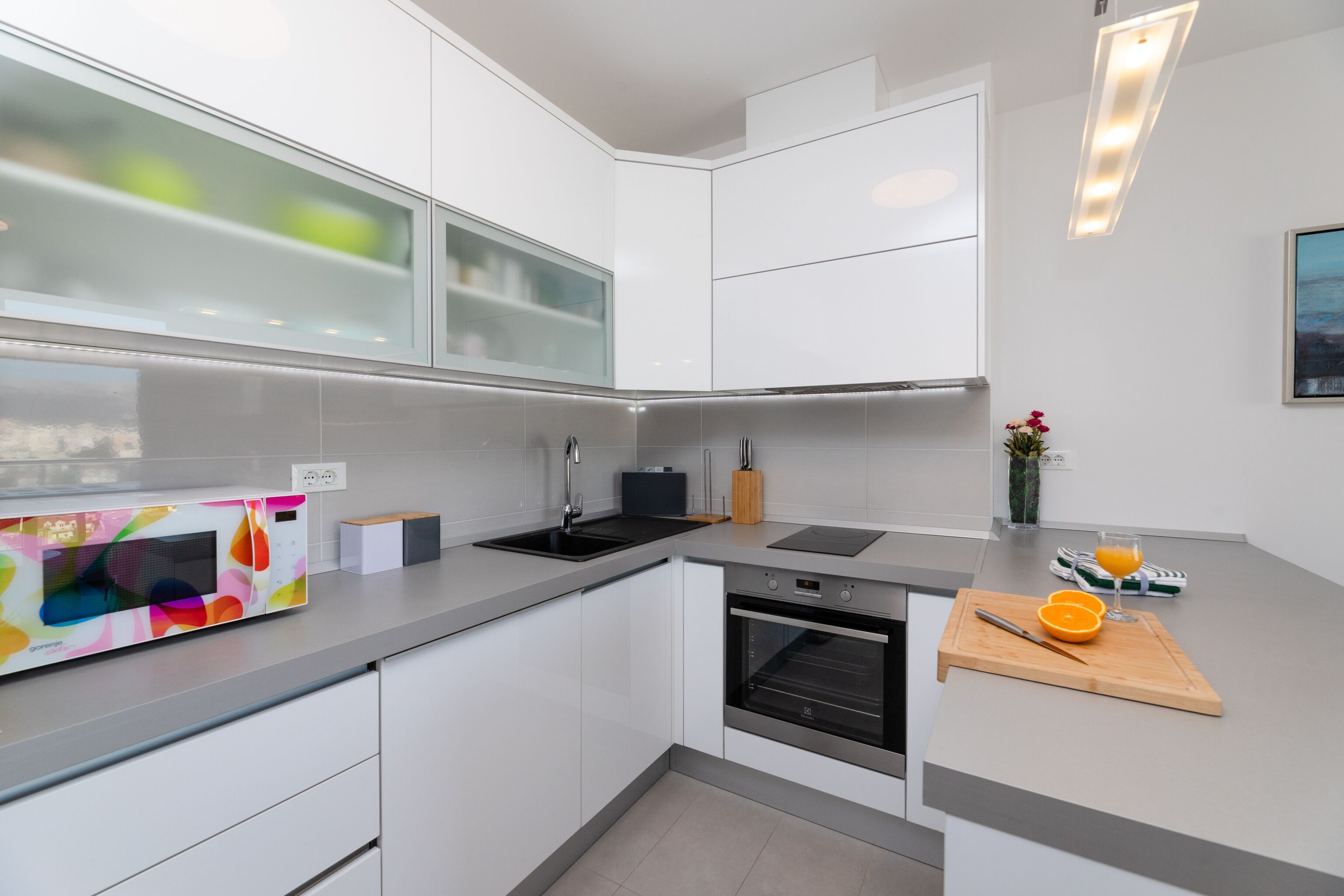 A modern kitchen with white cabinets, a built-in oven, a microwave, and a countertop with a cutting board and orange slices.
