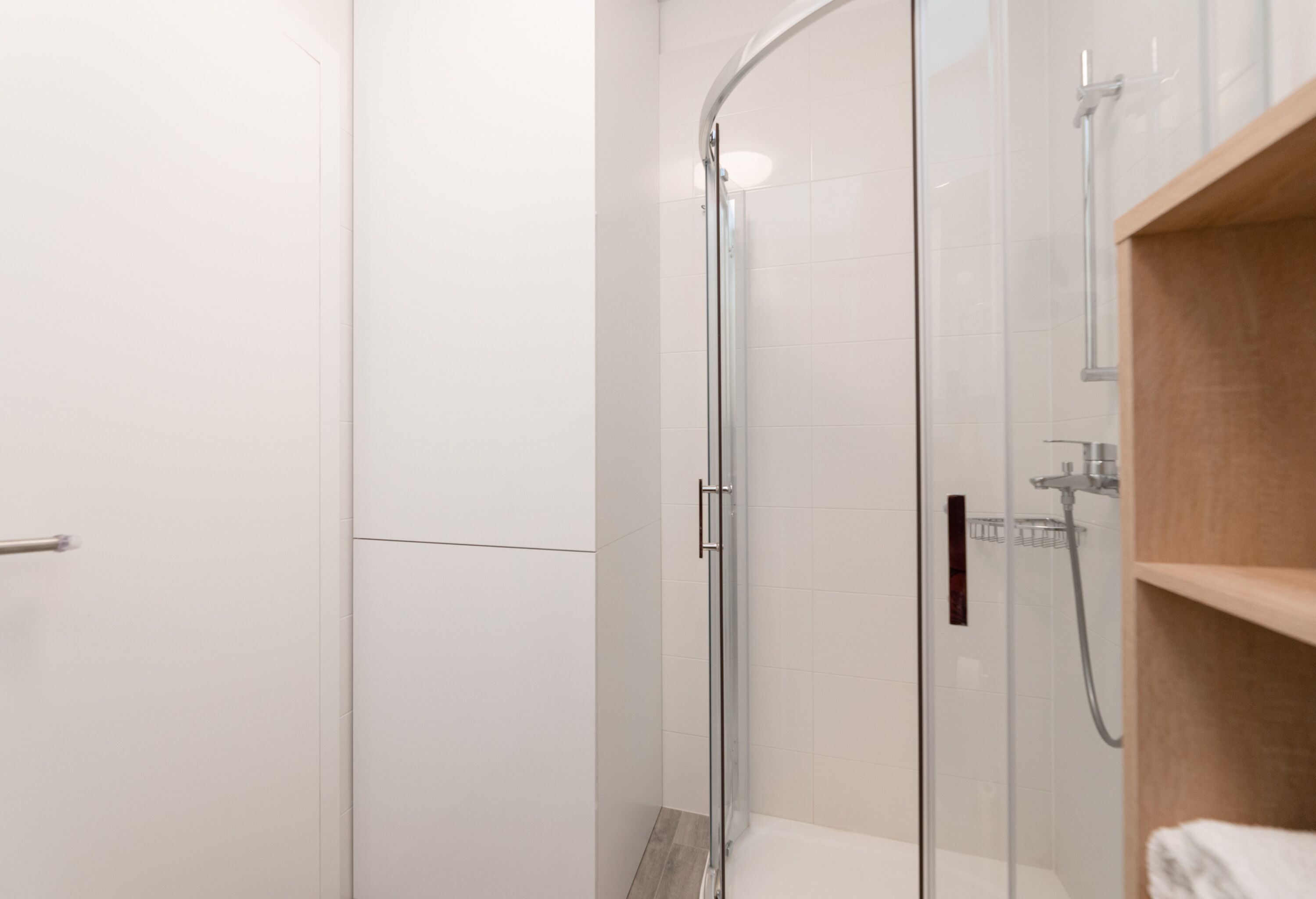 A modern bathroom with a glass shower enclosure, a wooden vanity, and a white tiled floor.