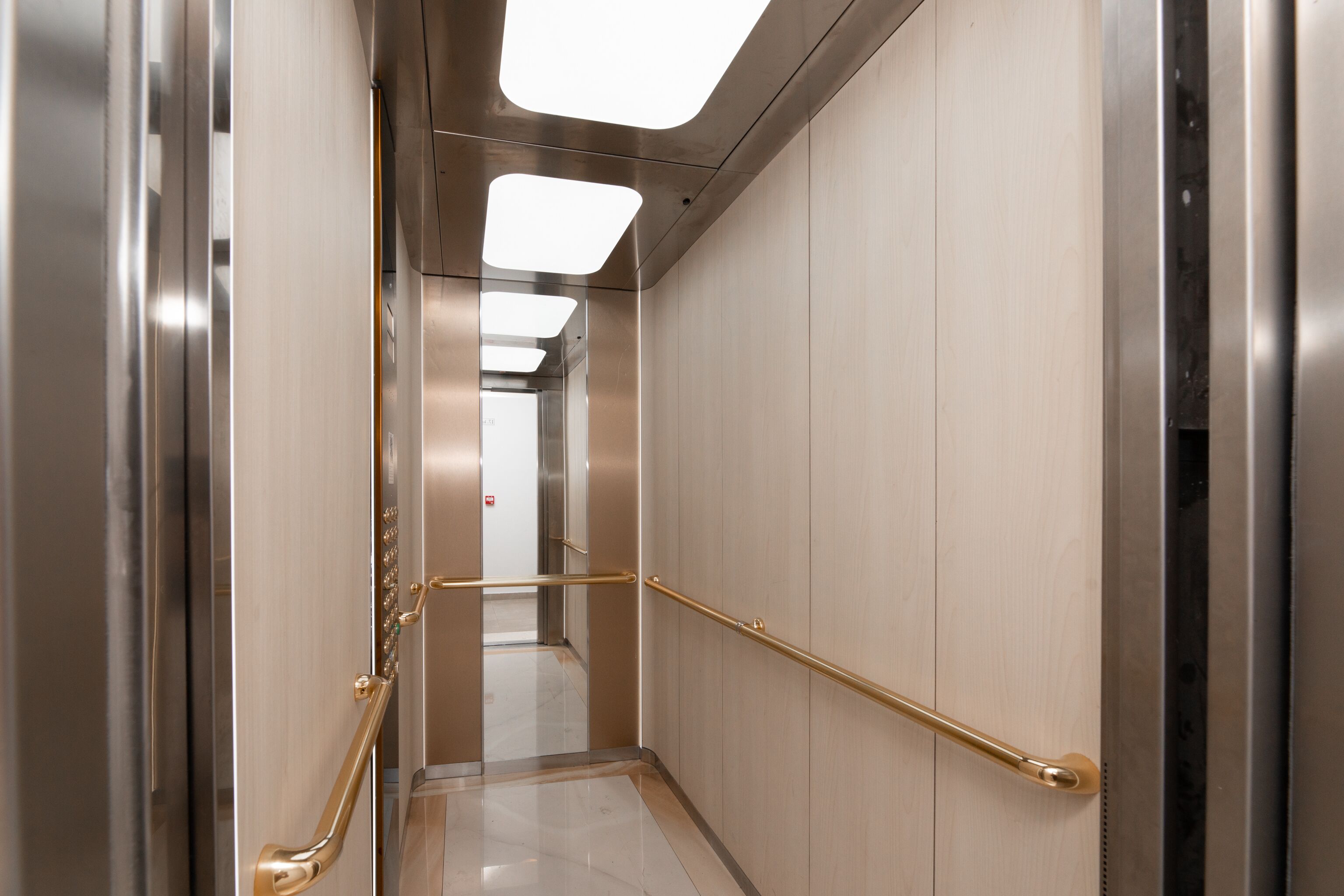 A modern elevator interior with metallic walls, a marble floor, and gold handrails.