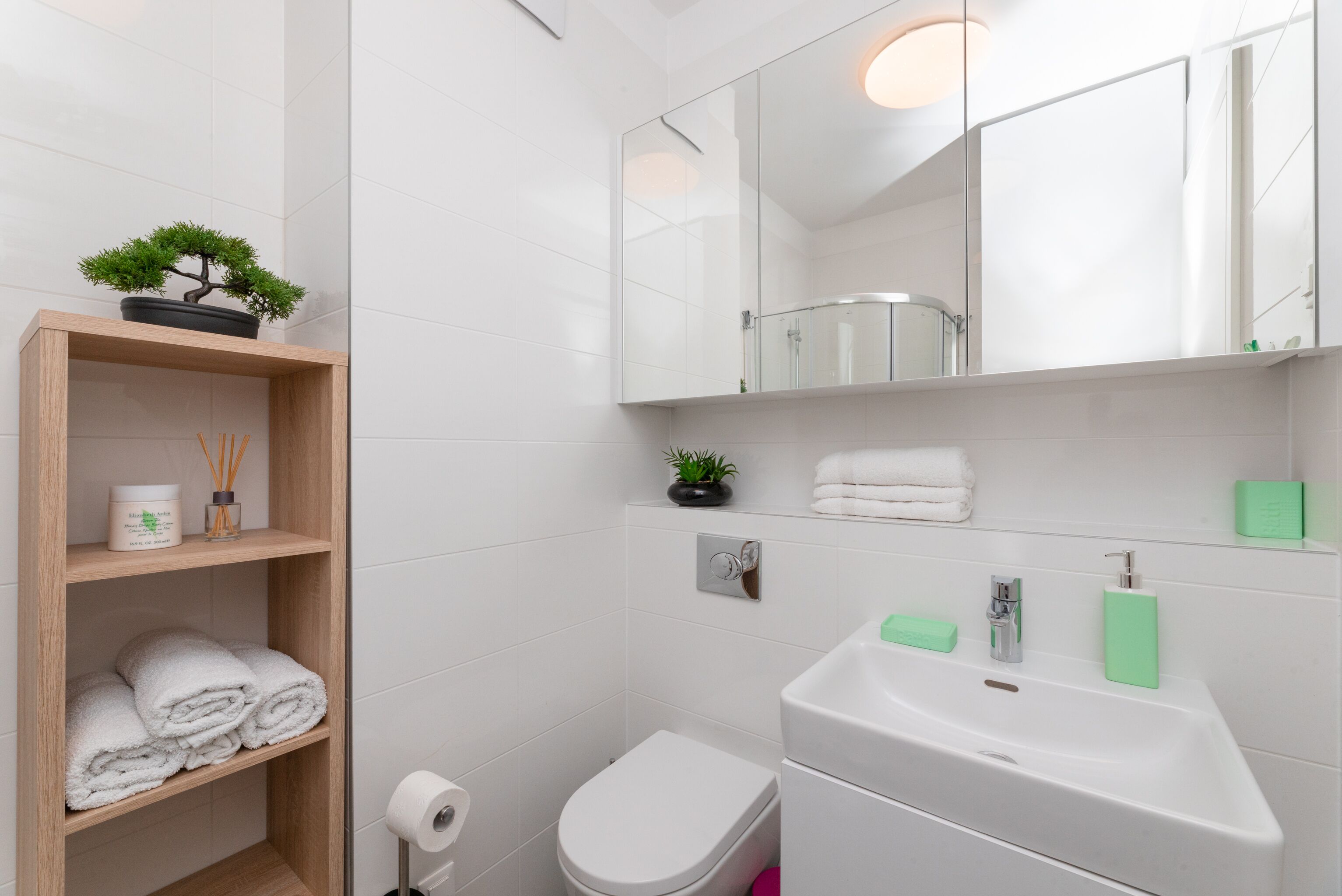 Et moderne badeværelse med toilet, vask og spejl. Der er en hylde med håndklæder og en lille potteplante.