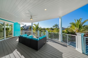 Une terrasse extérieure couverte, dotée d’un canapé en osier noir et de coussins turquoise, entourée d’une balustrade blanche et de palmiers.
