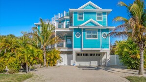 A two-story house with a garage, surrounded by palm trees and a clear blue sky. - Bluetique:  Monthly luxury beach home, private pool, rooftop deck (Holmes Beach)