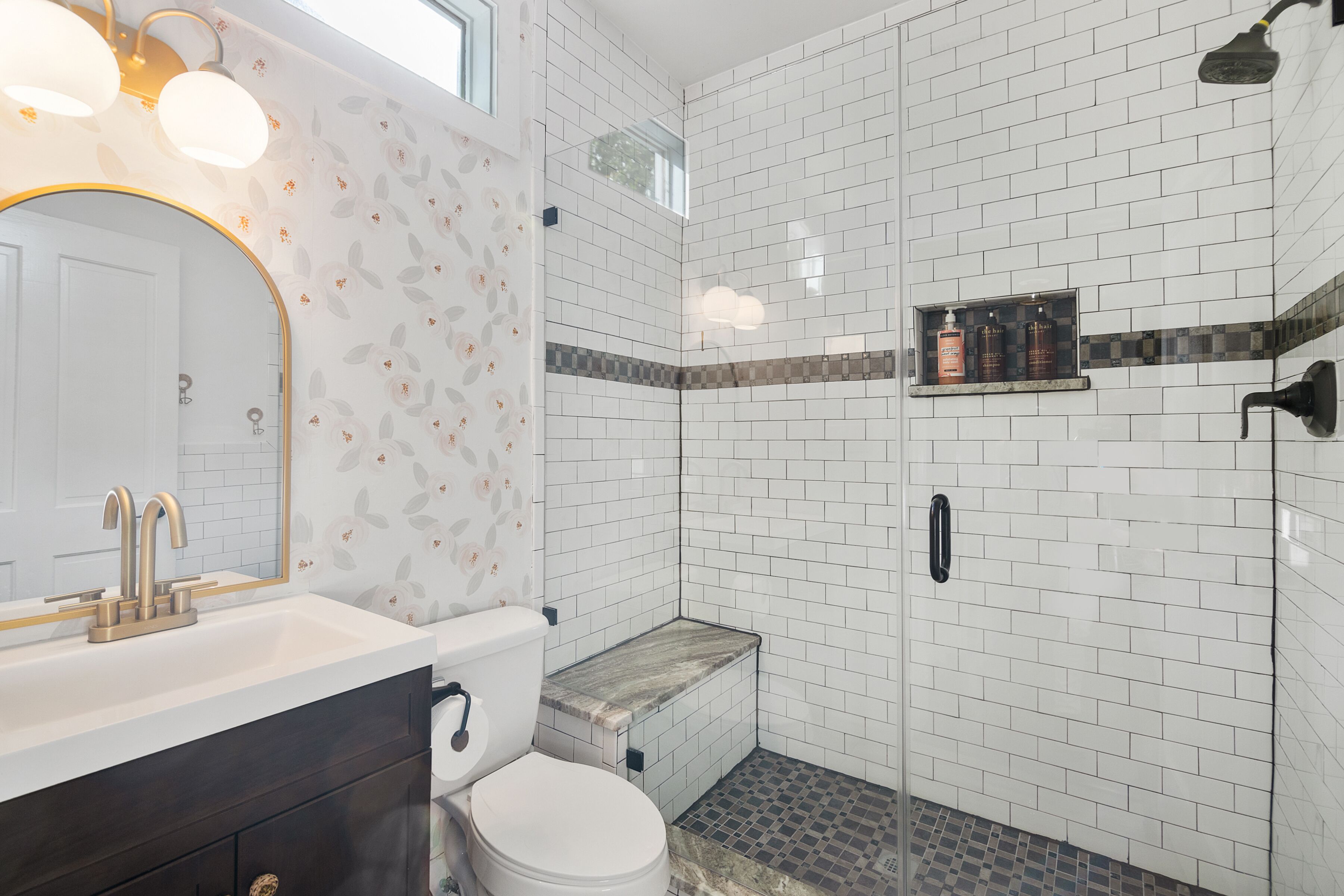 A bathroom with white tiled walls, a wooden vanity, a white toilet, and a shower area.