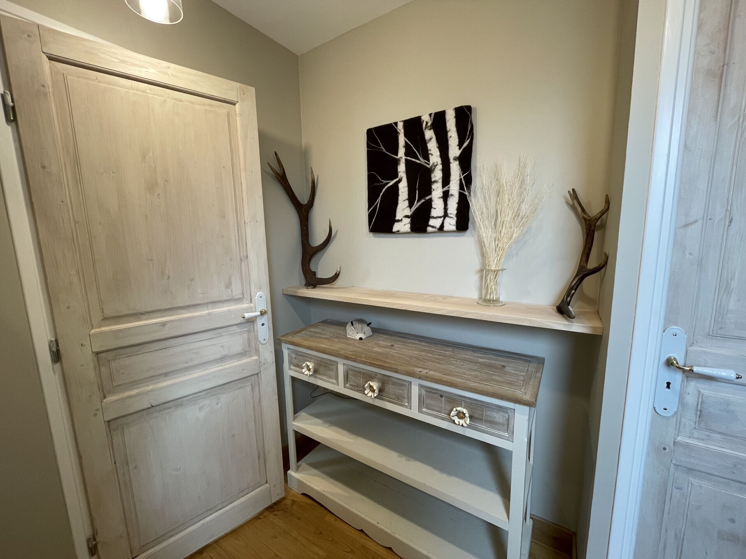 Een gang met een houten deur, een witte console tafel met lades, een aan de muur hangend schilderij en decoraties van gewei.