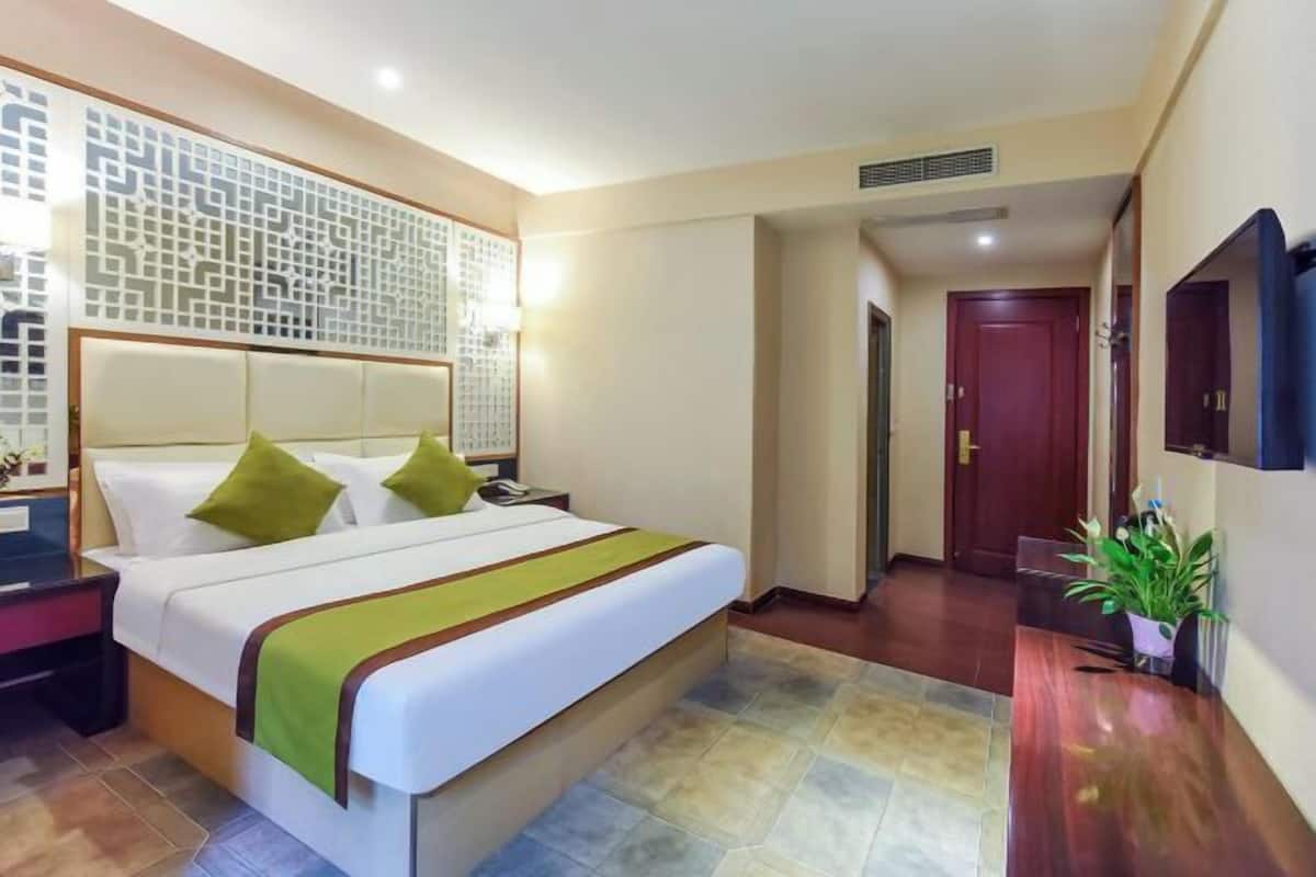A hotel room with a bed, bedside table, lamp, decorative wall panel, and a television mounted on the wall.