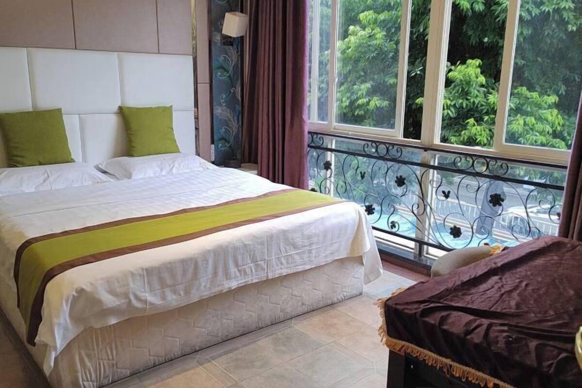A hotel room with a large bed, a view of greenery through the window, and a balcony with a wrought iron railing.