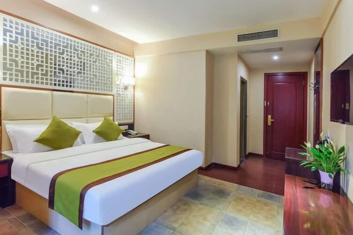 A hotel room with a bed, bedside table, decorative wall panel, and a wooden desk with a plant.