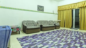 A living room with patterned sofas, a patterned rug, a table with a blue cover, and a sliding glass door with yellow curtains. - OYO Home 90492 Sena Homestay (Pokok Sena)