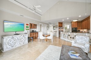 Un vaste espace de vie doté d’un grand téléviseur à écran plat, d’un ventilateur de plafond et d’un coin repas avec une table et des chaises blanches.