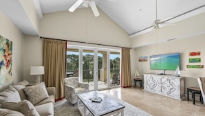 A spacious living room with a large sofa, a coffee table, and a television. There is a sliding glass door leading to a balcony with a view of the outdoors. - Golf Cart ~ Golf Course View ~ Community Pool (Miramar Beach)