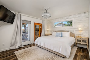 A bedroom with a large bed, a TV mounted on the wall, a wooden headboard, a window with a view, and a chandelier.