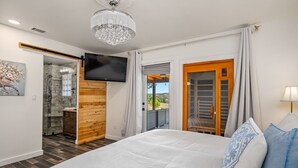 Un dormitorio con una cama grande, un televisor instalado en la pared, una puerta de madera que da a un balcón y una zona de ducha visible a través de una puerta abierta.