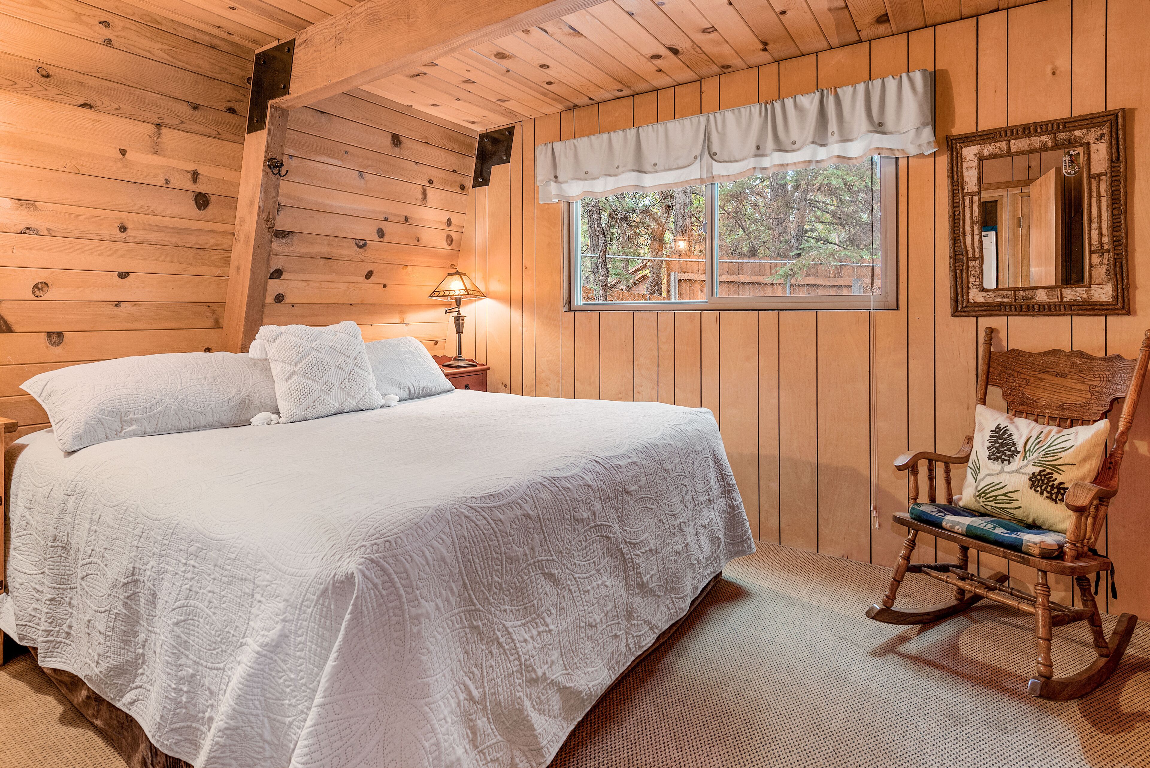 Un dormitorio de cabaña de madera con una cama grande, un sillón mecedor, una ventana con vista a árboles y un espejo en la pared.