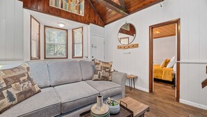 A cozy living room with a gray sofa, a wooden coffee table with decorative items, a mirror, and a door leading to a bedroom.