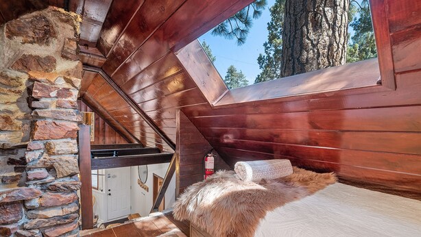 A cozy cabin bedroom with a stone wall, wooden ceiling, and a window overlooking trees.