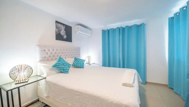 A bedroom with a white bed, blue curtains, a white headboard, and a wall-mounted air conditioner. - Fully Equipped New 3BR APT->DT->2Mins to the Beach (Sosúa)