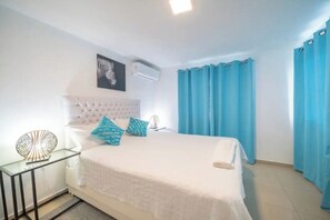 Een slaapkamer met een wit bed, blauwe gordijnen, een wit hoofdeinde en een wandairconditioner.