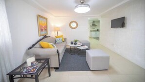 A modern living room with a sofa, coffee table, and ottoman. A flat-screen TV is mounted on the wall. - Fully Equipped New 3BR APT->DT->2Mins to the Beach (Sosúa)