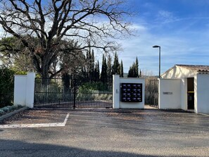 A gated parking area with a digital parking meter, a white building, and a large tree.