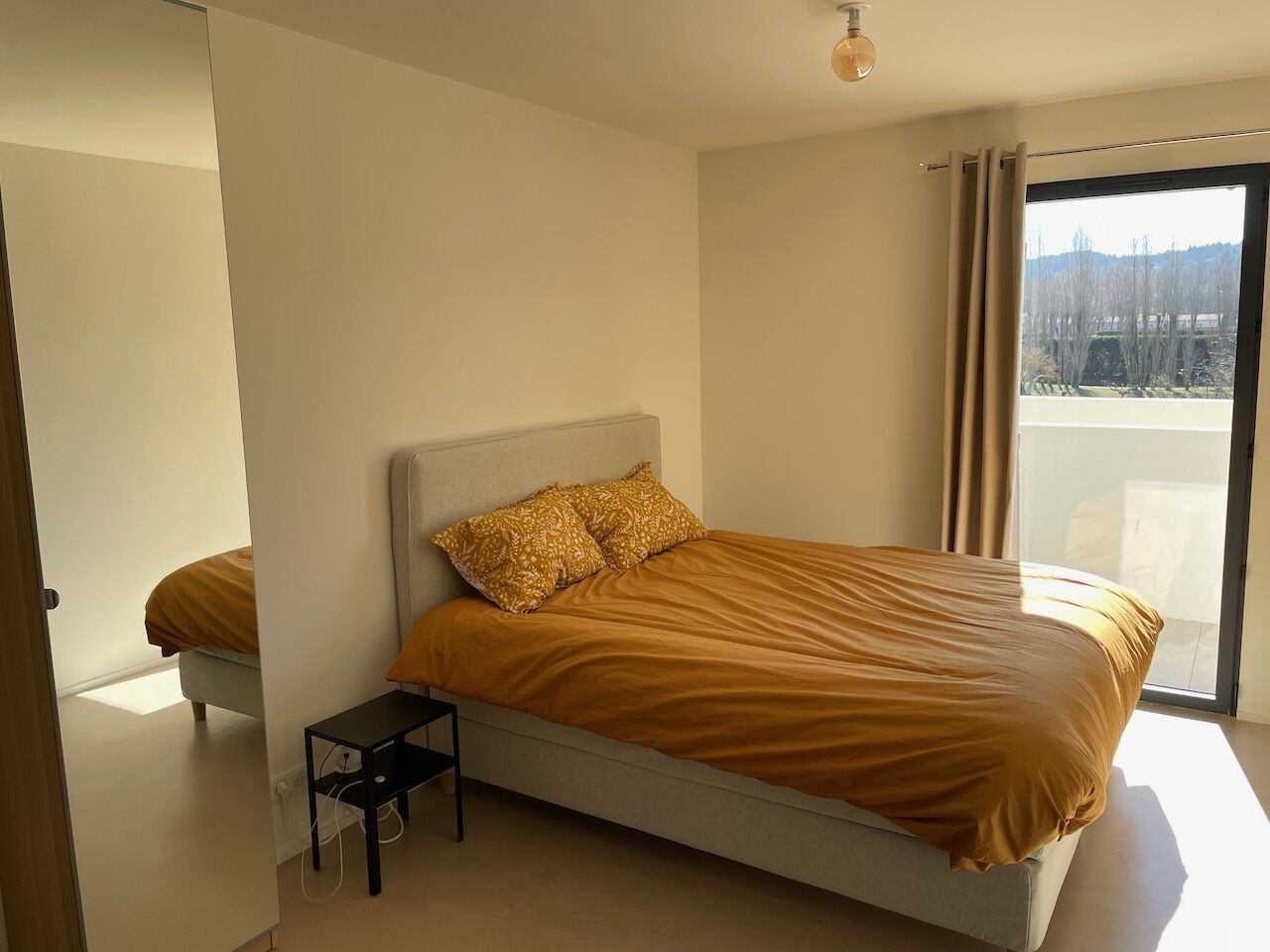 Een moderne slaapkamer met een groot bed, een nachtkastje en uitzicht op de omgeving door een groot raam.