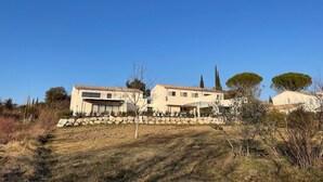 Una casa moderna de dos pisos con techo de tejas, rodeada de pasto seco y árboles dispersos.
