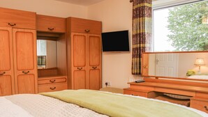 Ein Schlafzimmer mit Bett, Holzschrank, Flachbildfernseher, Spiegel und einem Fenster mit Vorhängen.