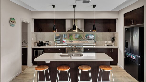 Una cocina moderna con una isla central, armarios oscuros, iluminación colgante y un refrigerador de acero inoxidable.