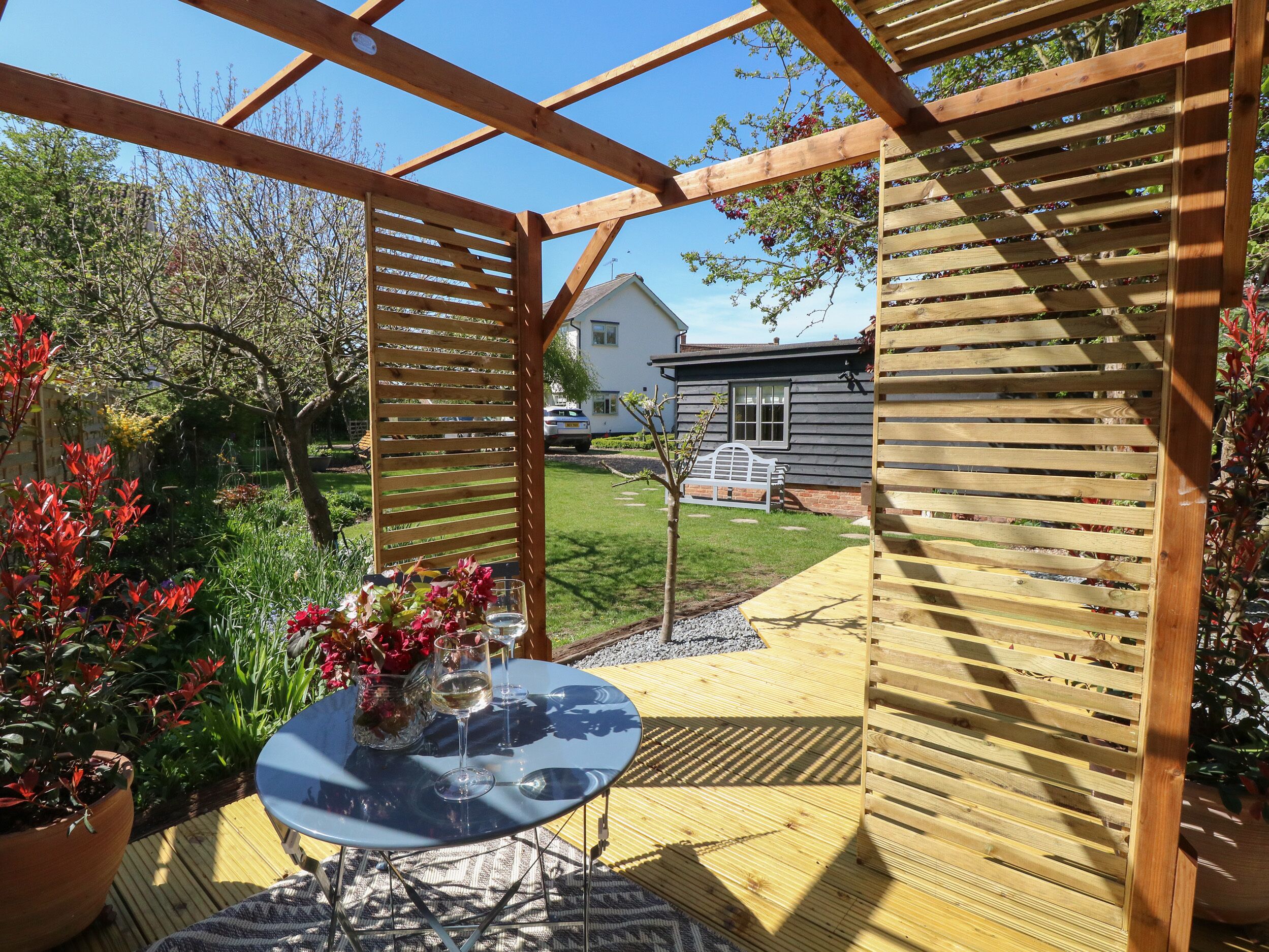 A wooden pergola with a glass table and chairs, surrounded by a garden with various plants and a bench.