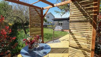 A wooden pergola with a glass table and chairs, surrounded by a garden with various plants and a bench.