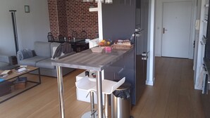 Ein modernes Wohnzimmer mit einem grauen Sofa, einem Holztisch und einer kleinen Küchenzeile mit einem grauen Schrank und einem Edelstahl-Mülleimer.