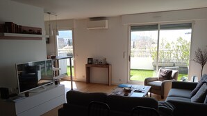 Ein modernes Wohnzimmer mit Flachbildfernseher, einer Couch, einem Couchtisch und einer Schiebetür zu einem Balkon mit Blick auf die Stadt.