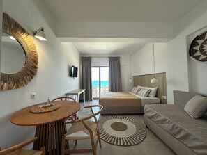 A modern hotel room with a bed, a sofa, a dining area with a table and chairs, a TV, and a view of the sea through a large window.