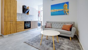 A modern kitchen with wooden cabinets, a built-in oven, and a washing machine. A grey sofa with two cushions is in the living area, and a painting of a coastal scene hangs on the wall. - Perfect Location, Idyllic 2BR Apartment (Sliema)
