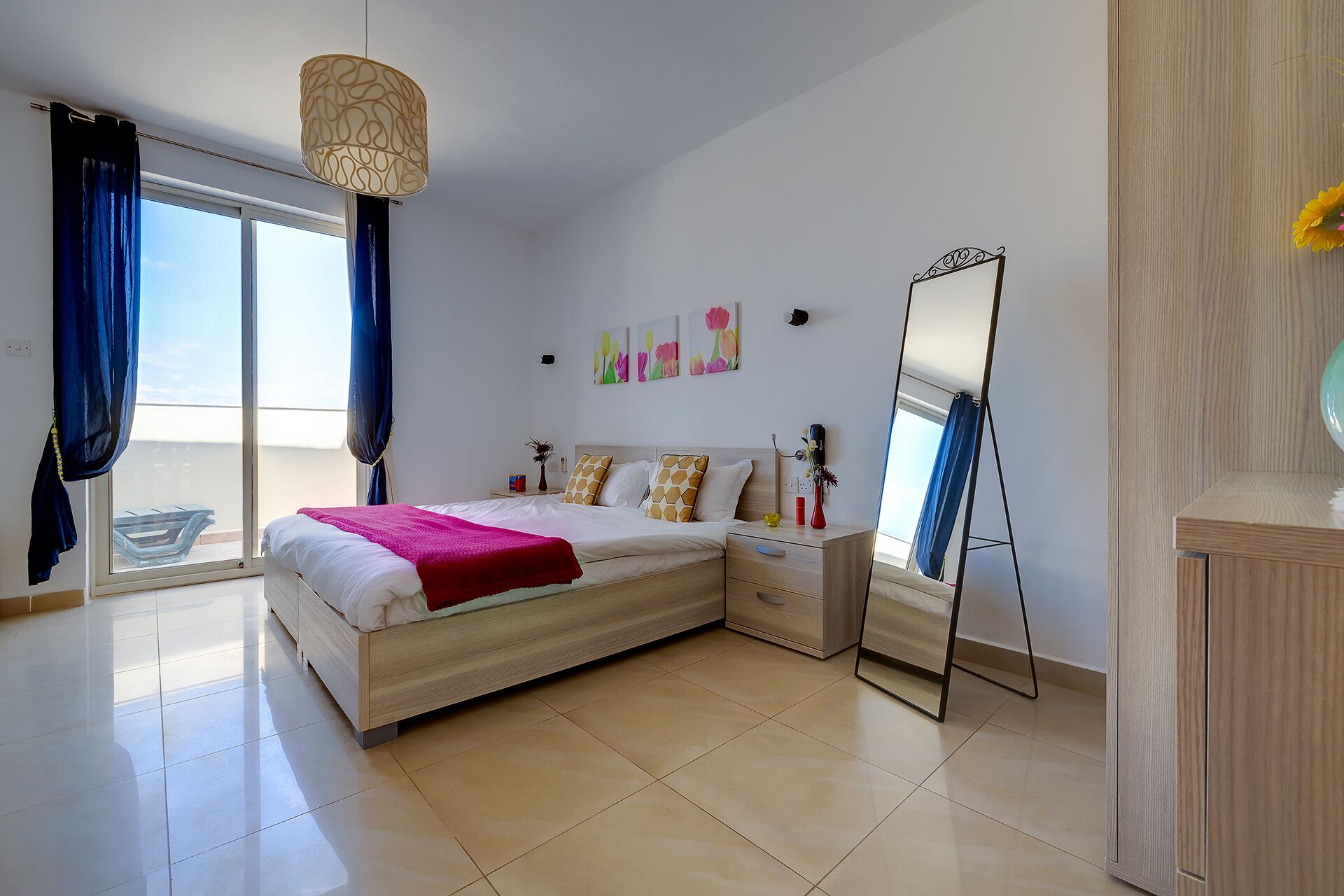 A modern bedroom with a large bed, a bedside table, a mirror, and a view of the outdoors.