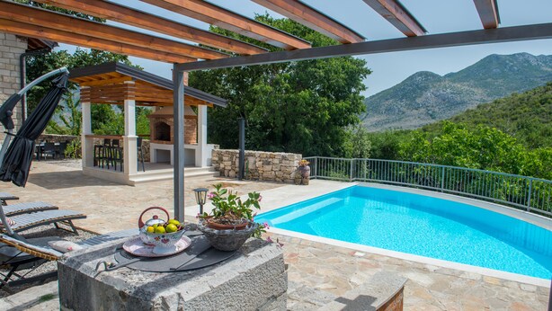 Una piscina con terraza de piedra, una pérgola, mobiliario exterior y vistas a la montaña.