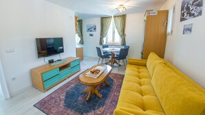 Una sala de estar con un sofá amarillo, un televisor sobre un mueble de madera, una mesa de comedor con sillas y una alfombra estampada.