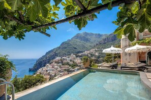 Vista a la piscina con vistas a un pueblo costero, edificios en una ladera, cielo azul despejado y montañas al fondo.