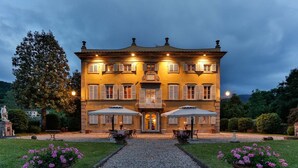 A large, two-story building with a symmetrical facade, multiple windows with shutters, and a central entrance with a balcony. There are outdoor seating areas with umbrellas and potted plants. - VILLA CONTE - Seven Bedroom Villa, Sleeps 14 (Lucca)
