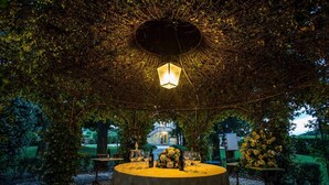 An outdoor dining area with a round table set for a meal, surrounded by greenery and a decorative lantern overhead. - VILLA CONTE - Seven Bedroom Villa, Sleeps 14 (Lucca)