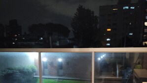 A balcony with a glass railing, a tiled floor, and a view of a building with lit windows. - Modern, cosy and practical place (Montevideo)