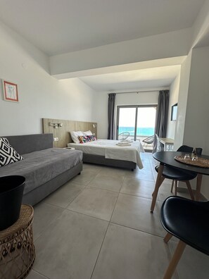 A modern hotel room with a bed, a sofa, a dining table, and a view of the sea.