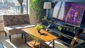 Ein modernes Wohnzimmer mit einer Couch, einem Couchtisch aus Holz, einem schwarzen Ledersofa, einem Fernseher und Blick auf eine Stadtlandschaft durch große Fenster.