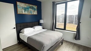 Ein modernes Schlafzimmer mit einem Bett, einem Gemälde, einem Fenster mit Vorhängen und Blick auf Gebäude.