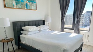 A modern bedroom with a grey upholstered bed, white linens, a round bedside table with a lamp, and a large window with curtains. - 2BR apt close to EWR Airport m5og (Elizabeth)
