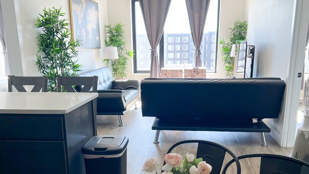 A modern living room with a glass dining table, a sofa, and a view of buildings through large windows. - 2BR apt close to EWR Airport m5og (Elizabeth)