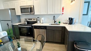 A modern kitchen with white cabinets, stainless steel appliances, a glass dining table, and a black trash bin. - 2BR apt close to EWR Airport m5og (Elizabeth)